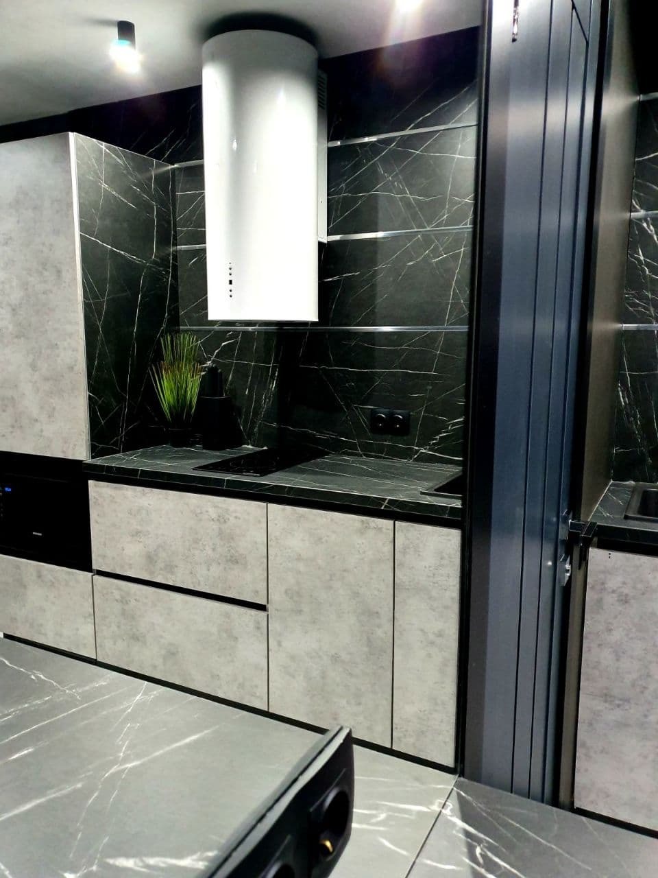 Nowoczesna kuchnia z szarymi szafkami o fakturze betonu, czarnym blatem i ścianą z płytek imitujących czarny marmur z białymi żyłami. Na blacie roślina w czarnej doniczce, nad blatem biały...