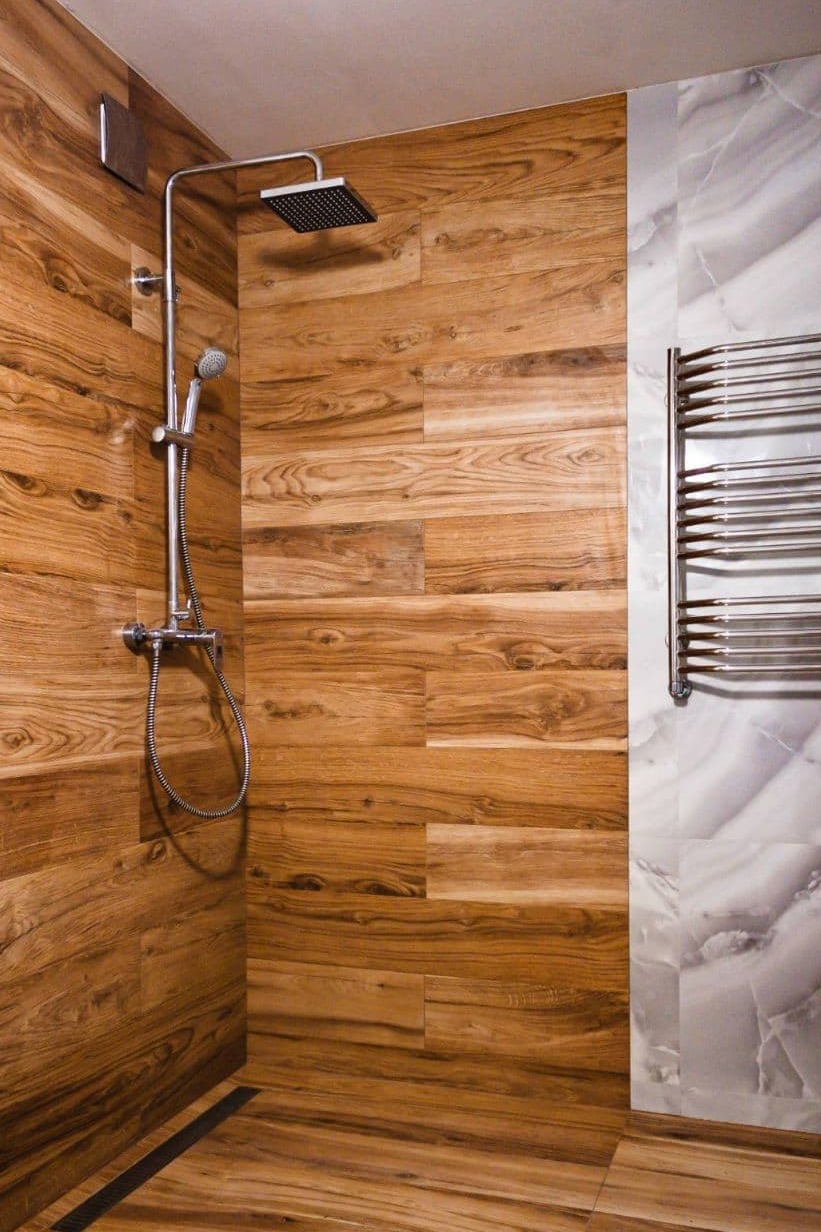 Kabina prysznicowa wyłożona płytkami imitującymi drewno, z chromowaną armaturą i grzejnikiem drabinkowym na tle marmurowych płytek.