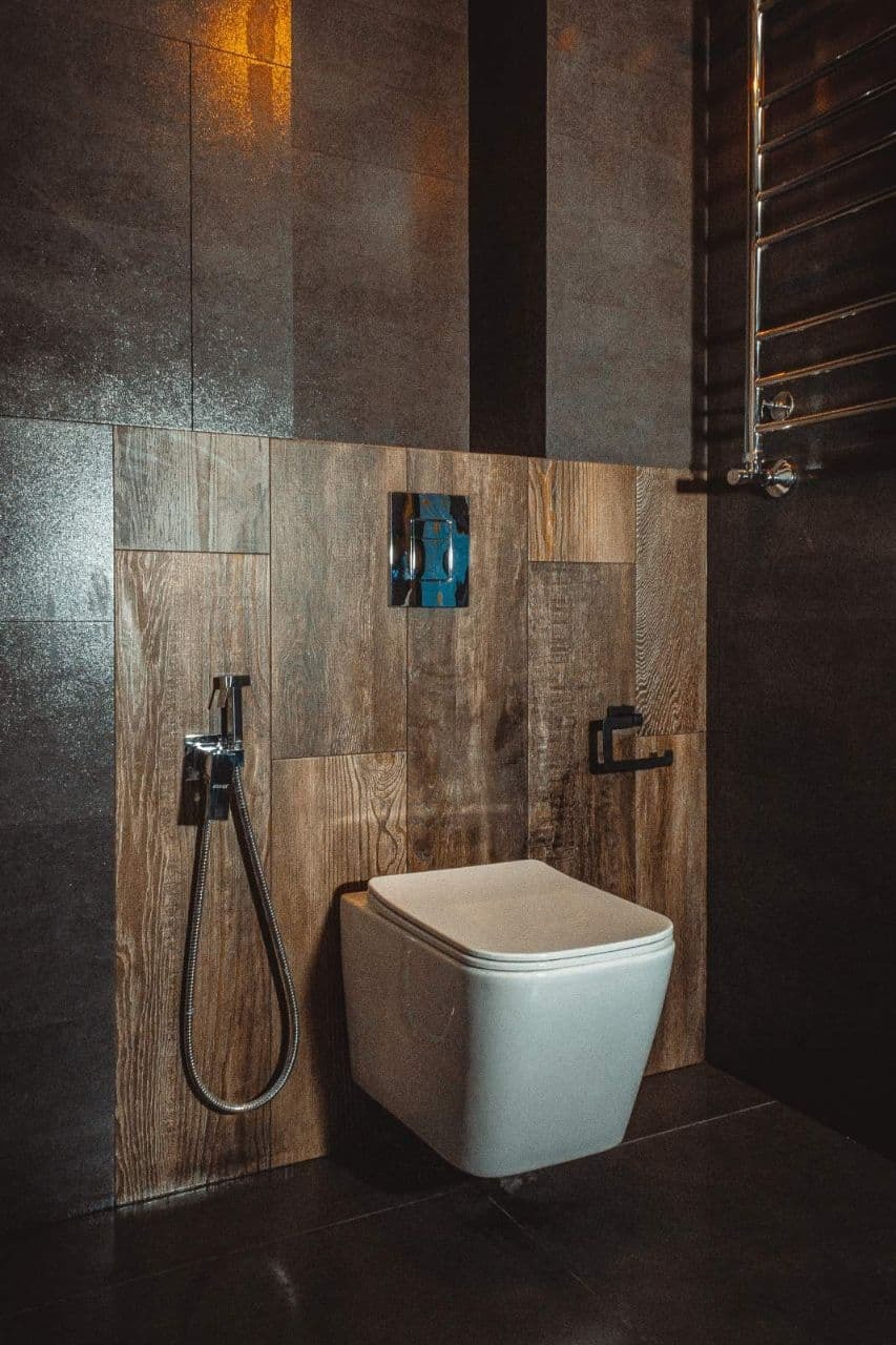 Nowoczesna toaleta podwieszana na ścianie wyłożonej płytkami imitującymi drewno, z ciemnymi płytkami na pozostałych ścianach i podłodze, oraz chromowaną armaturą i grzejnikiem drabinkowym.
