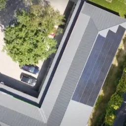 Widok z góry na dom z panelami słonecznymi na dachu, obok zaparkowane samochody, zadbany ogród i kamienny podjazd.