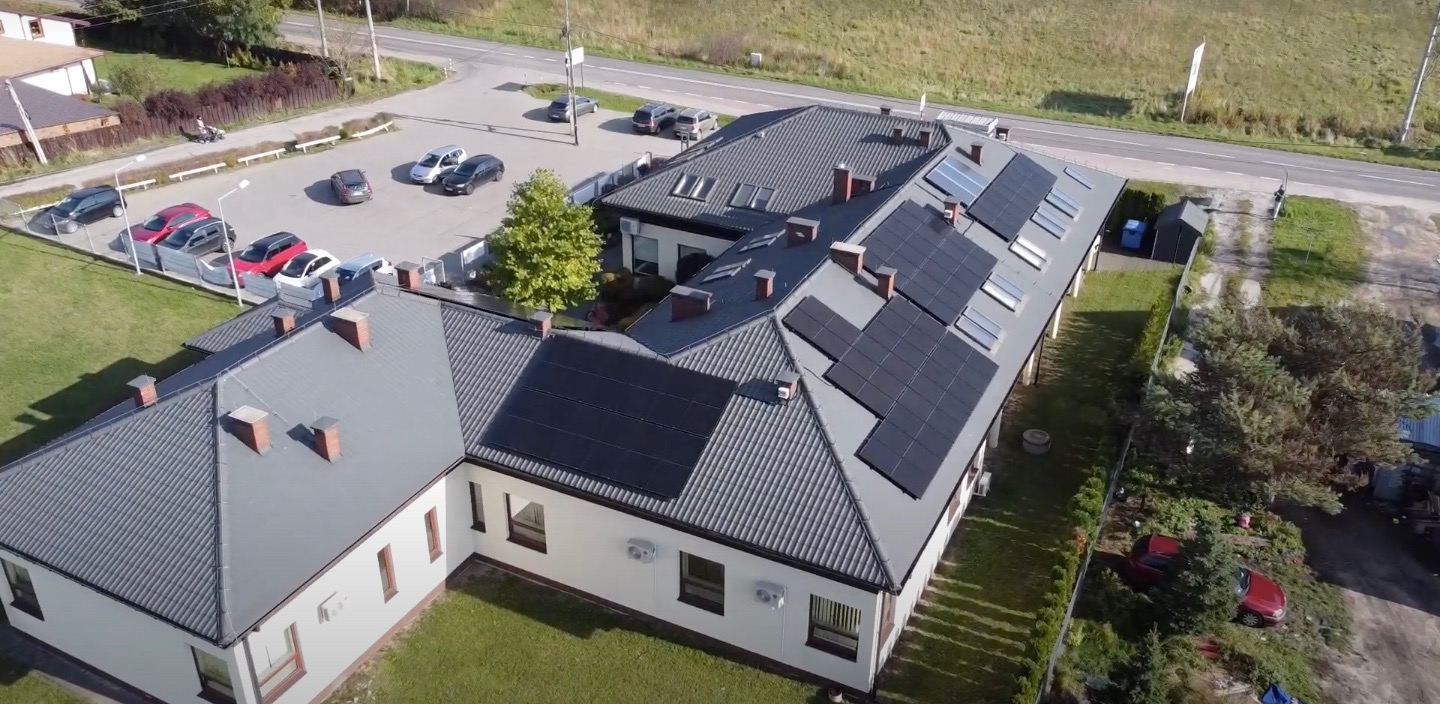Widok z góry na budynek z szarym dachem i panelami słonecznymi, parking z samochodami i teren zielony