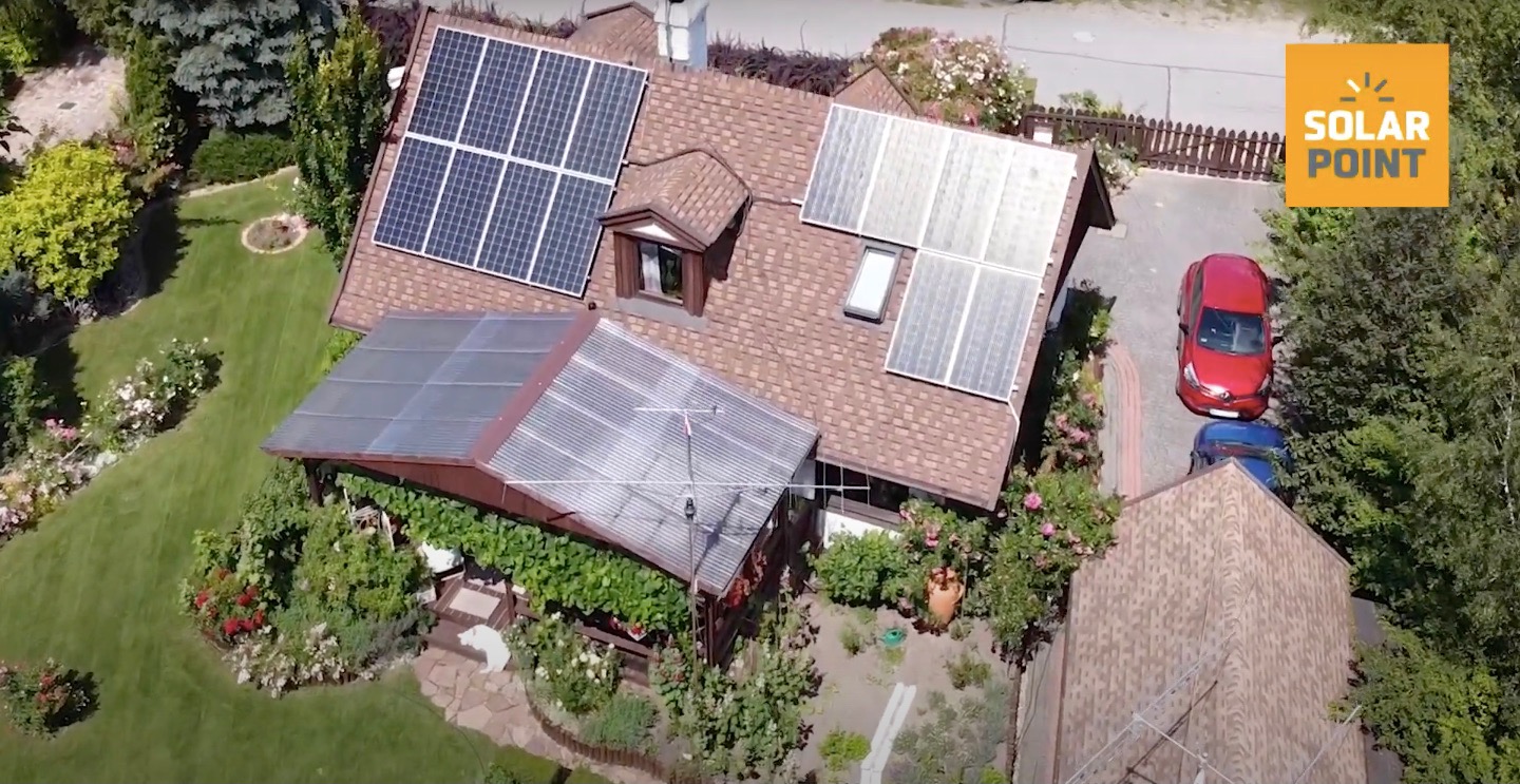 Widok z drona na dom z panelami słonecznymi na dachu, zadbany ogród, dwa samochody na podjeździe i logo firmy Solar Point w prawym górnym rogu.