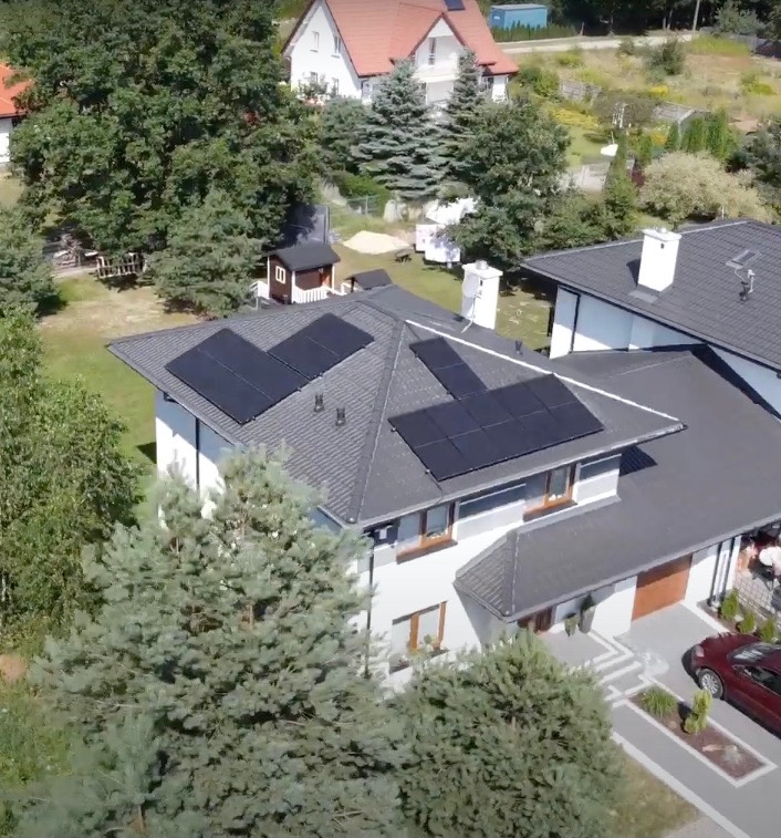 Dom jednorodzinny z szarą dachówką i panelami fotowoltaicznymi ułożonymi w dwóch grupach, widok z góry, otoczony zielenią i innymi budynkami.