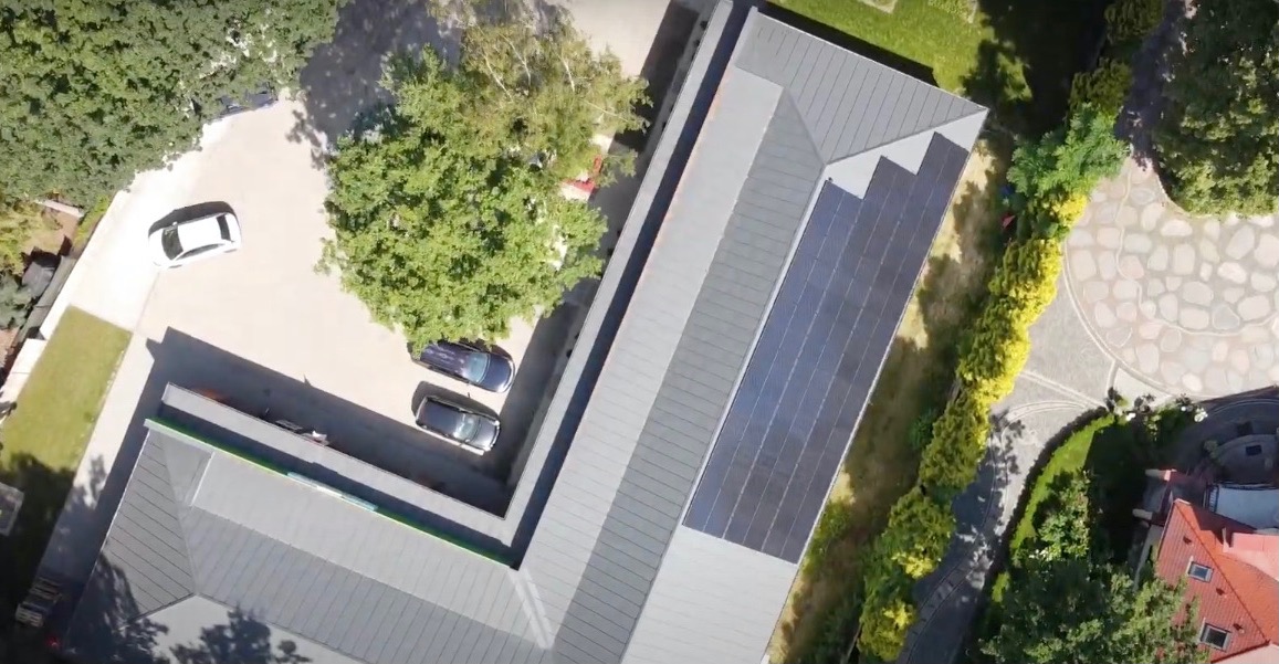 Widok z góry na dom z panelami słonecznymi na dachu, obok zaparkowane samochody, zadbany ogród i kamienny podjazd.