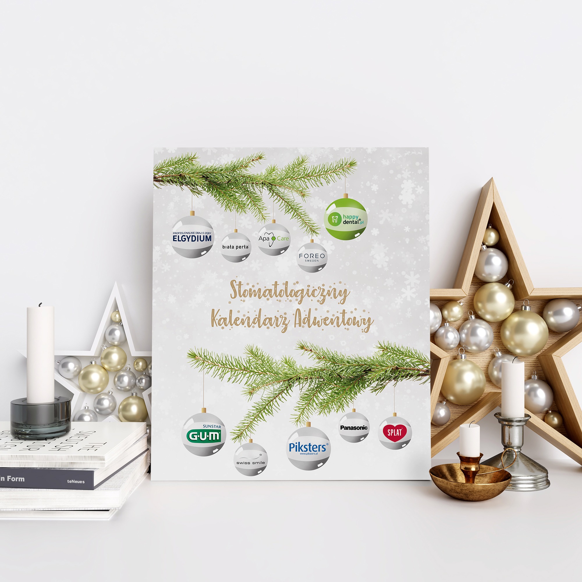 Stomatologiczny kalendarz adwentowy z logotypami firm dentystycznych zawieszonymi na gałązkach choinki, ustawiony na białym stole z książkami i dekoracjami świątecznymi.