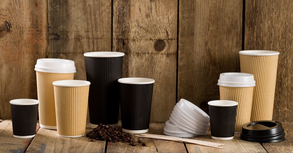Różnej wielkości papierowe kubki w kolorach beżowym i czarnym, z wieczkami i bez, ułożone na drewnianym blacie z ziarnami kawy i mieszadełkami, na tle drewnianej ściany.