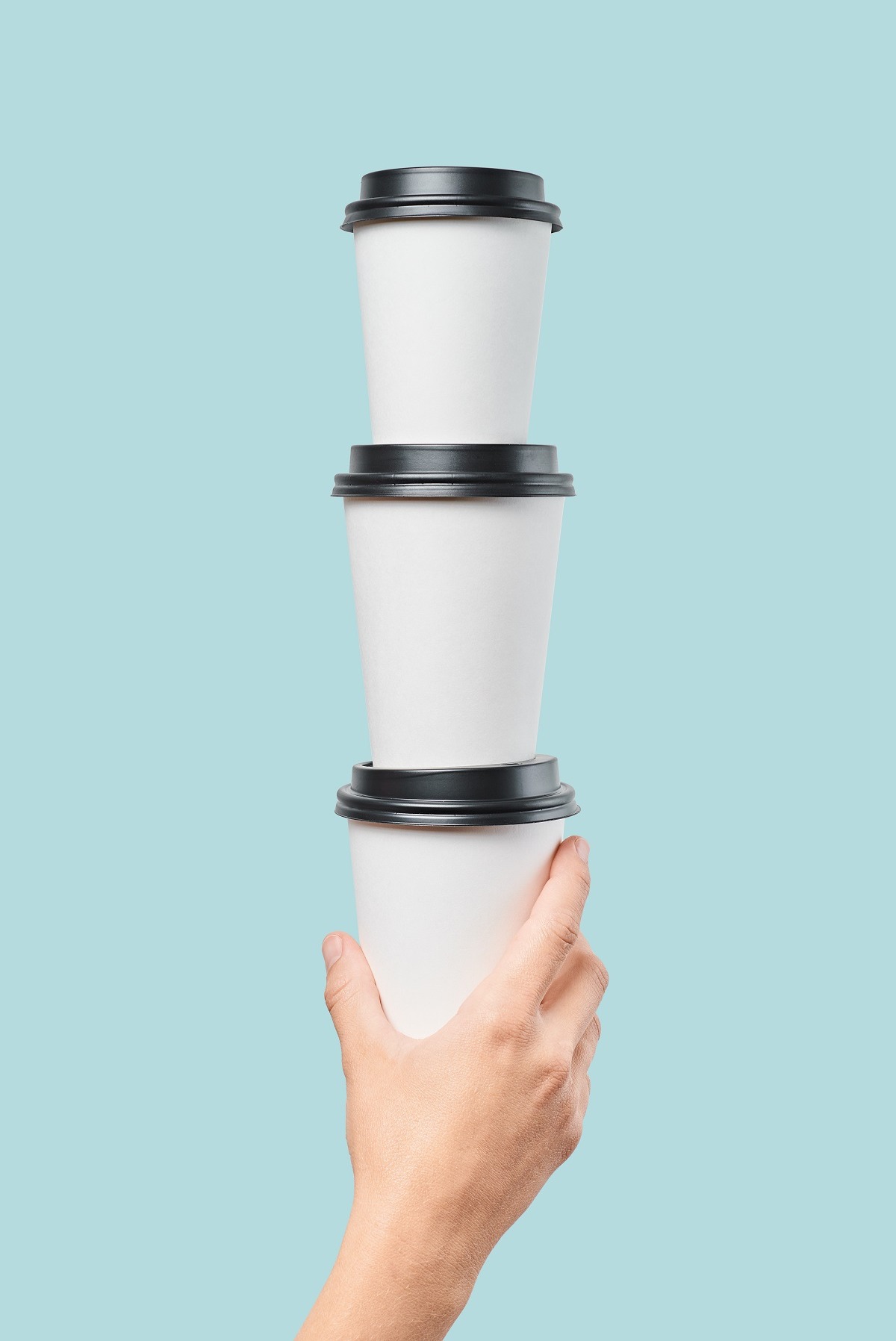 Dłoń trzymająca w pionie trzy białe, papierowe kubki z czarnymi pokrywkami na tle błękitnego tła.