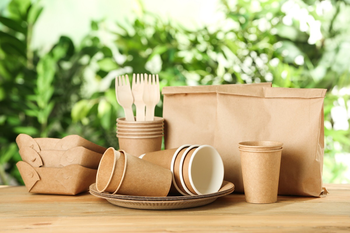 Zestaw ekologicznych naczyń jednorazowych: papierowe kubki, talerze, pudełka i drewniane sztućce na drewnianym stole, z rozmytym zielonym tłem roślinnym.