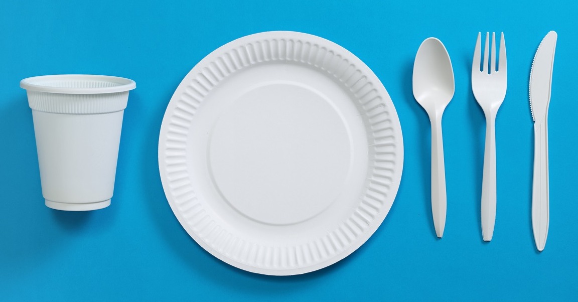 Zestaw jednorazowych naczyń: biały talerz papierowy, kubek z tworzywa, plastikowa łyżka, widelec i nóż, ułożone na niebieskim tle.