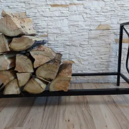 Czarny, metalowy stojak na drewno kominkowe, wypełniony połupanymi kawałkami drewna, ustawiony na drewnianej podłodze na tle ściany wyłożonej dekoracyjnym kamieniem.