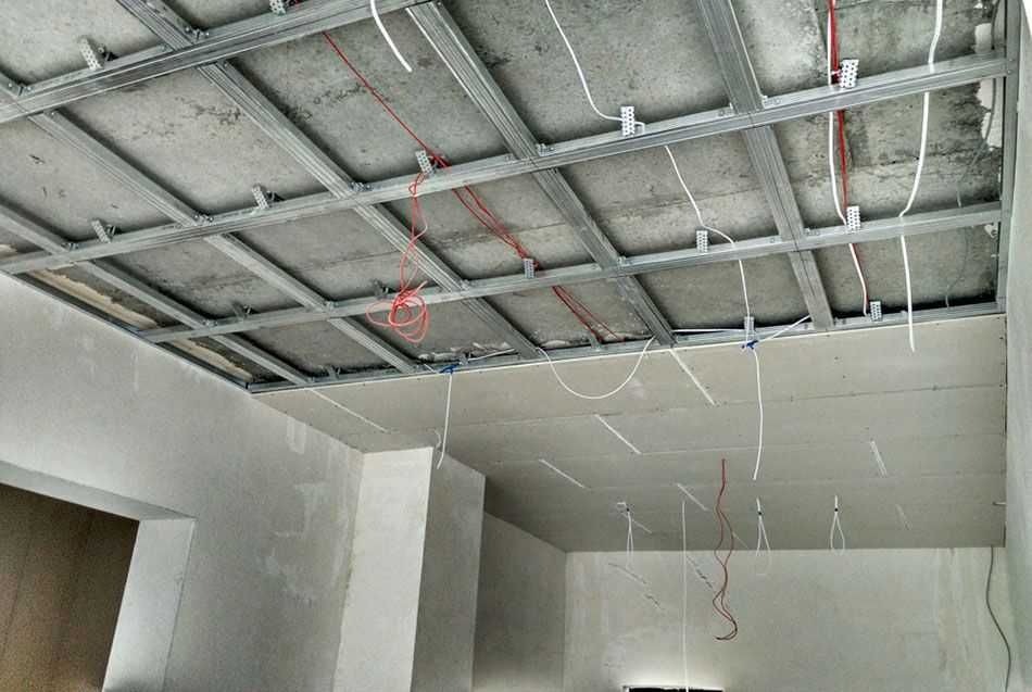 Stelaż sufitowy z profili metalowych, widoczne przewody elektryczne, częściowo zamontowane płyty na suficie, ściany w surowym tynku.