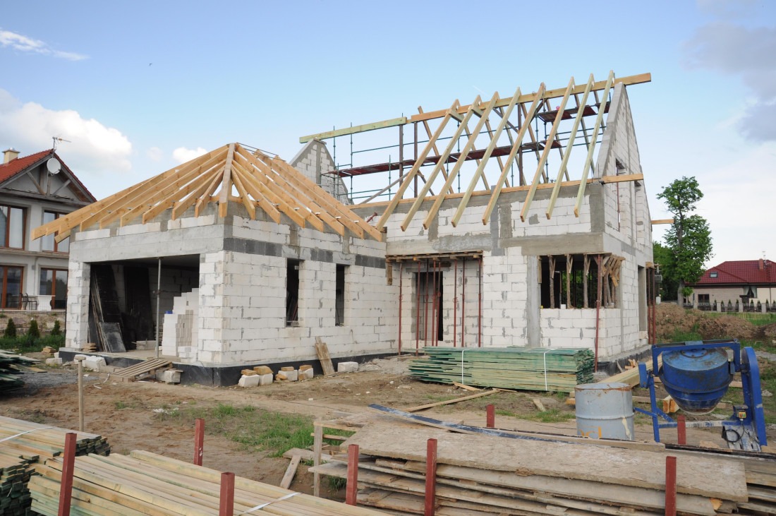 Budowa domu jednorodzinnego z białych bloczków, widoczne elementy konstrukcji dachu z drewna, rusztowania i betoniarka.