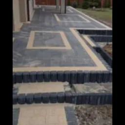 Ścieżka i schody z kostki brukowej w dwóch odcieniach szarości i beżu, z widocznymi obrzeżami, prowadzące wzdłuż budynku z ogrodem w tle.