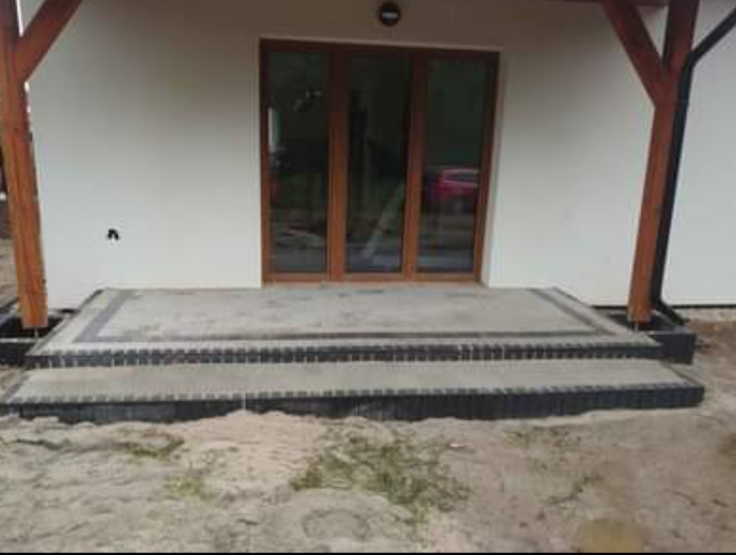 Wykończone dwustopniowe schody wejściowe do domu, obłożone szarą kostką brukową z ciemnymi obrzeżami, widoczne drewniane słupy werandy i częściowo brudne podłoże.