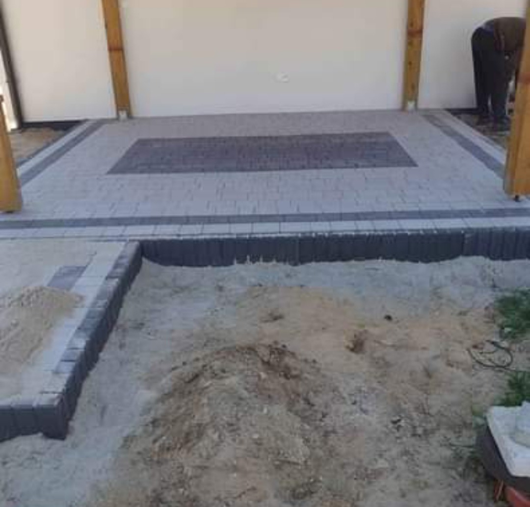 Fragment układanej nawierzchni z kostki brukowej w odcieniach szarości, widoczny obszar przygotowany pod dalsze prace ziemne z piaskiem i fragmentem zieleni, częściowo zadaszona przestrzeń...