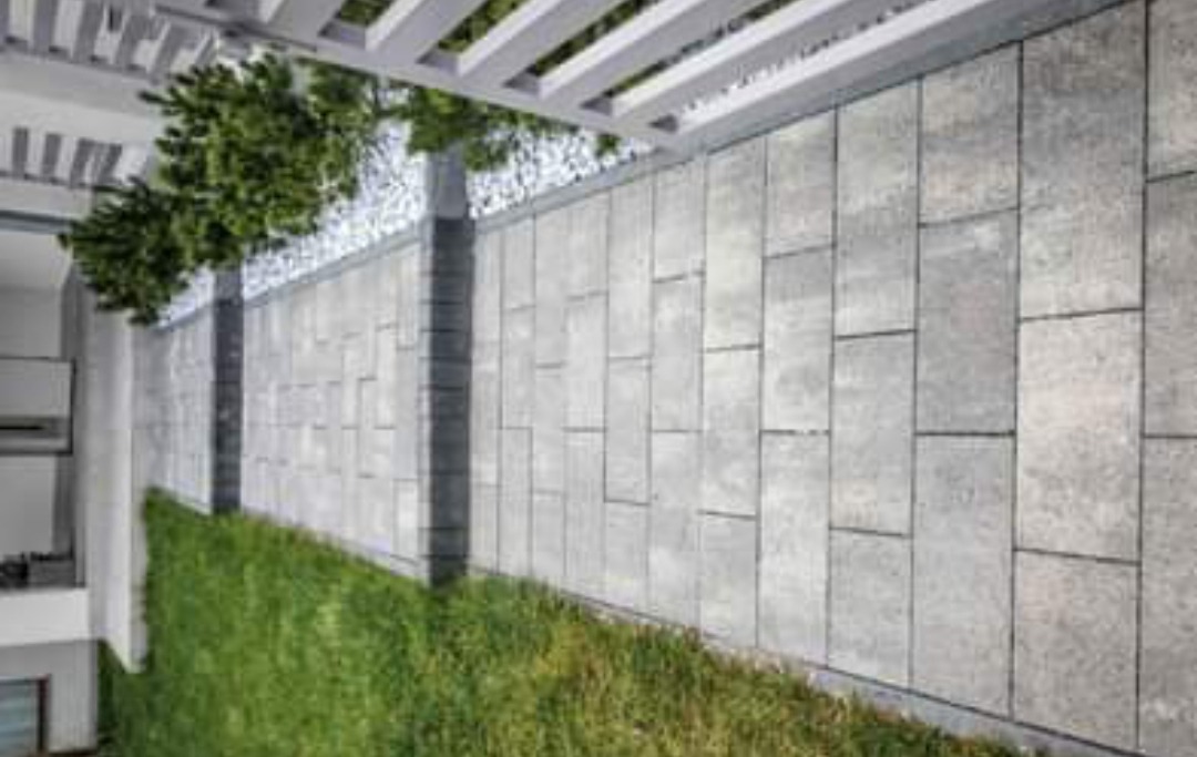 Nowoczesny taras wyłożony szarą kostką o nieregularnym kształcie, obok zielony trawnik i małe drzewka, całość zadaszona pergolą.