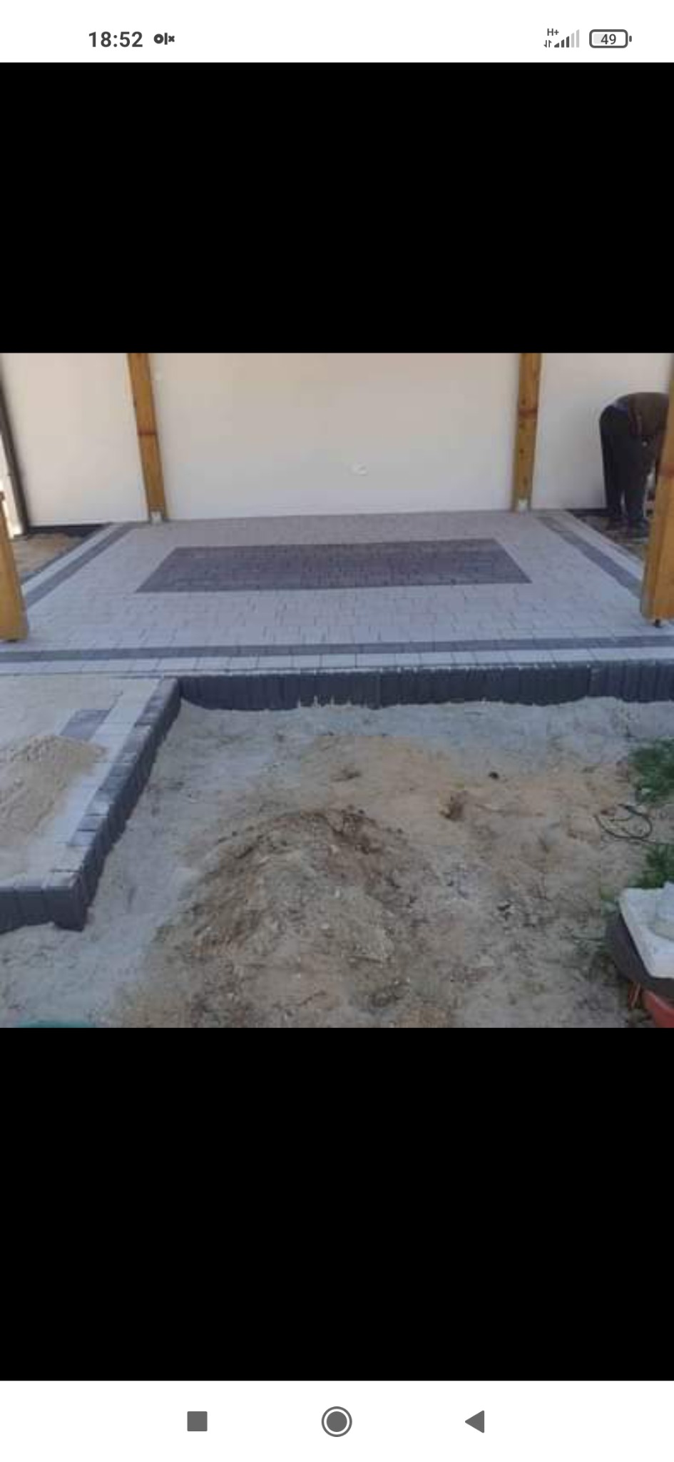 Fragment podwórza z częściowo ułożoną kostką brukową w odcieniach szarości, widoczny piasek i ziemia przygotowane pod dalsze prace, drewniane słupy konstrukcyjne w tle.