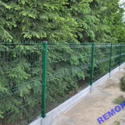 Zielone ogrodzenie z paneli metalowych na betonowej podmurówce, z widocznymi świerkami w tle.