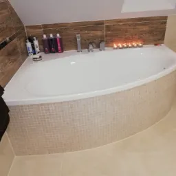 Narożna wanna obudowana mozaiką w beżowych odcieniach, armatura chromowana, na wannie zapalone świeczki, na półce kosmetyki i mydelniczka, ściany wyłożone płytkami imitującymi drewno i jasnymi...