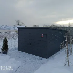 Grafitowy garaż z blachy falistej na tle zimowego krajobrazu, z małymi drzewkami iglastymi i ośnieżonymi budynkami w oddali.