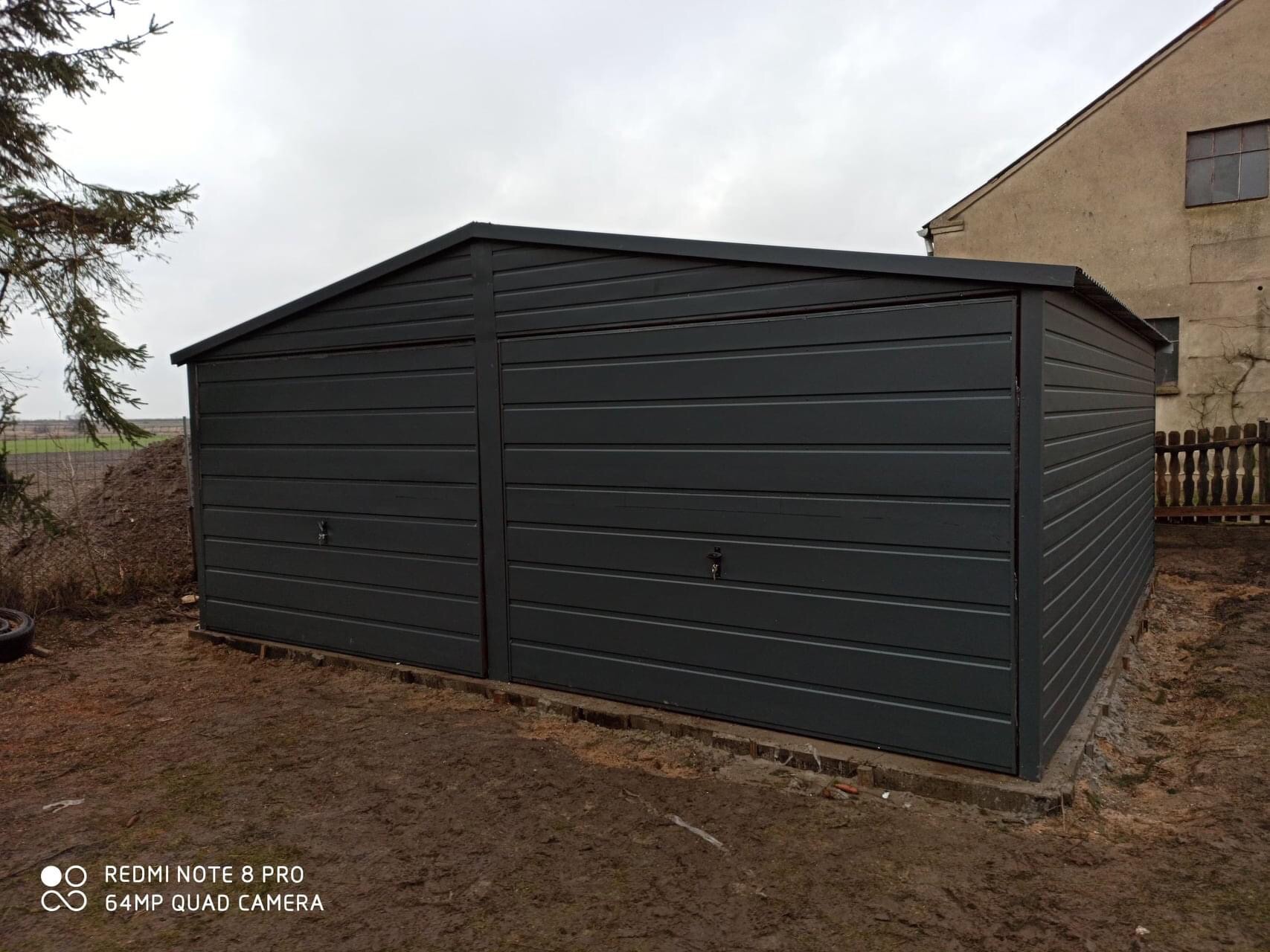 Grafitowy garaż blaszany dwustanowiskowy z dachem dwuspadowym na posesji w Przechlewie, widoczny z boku, na ziemi ślady błota.