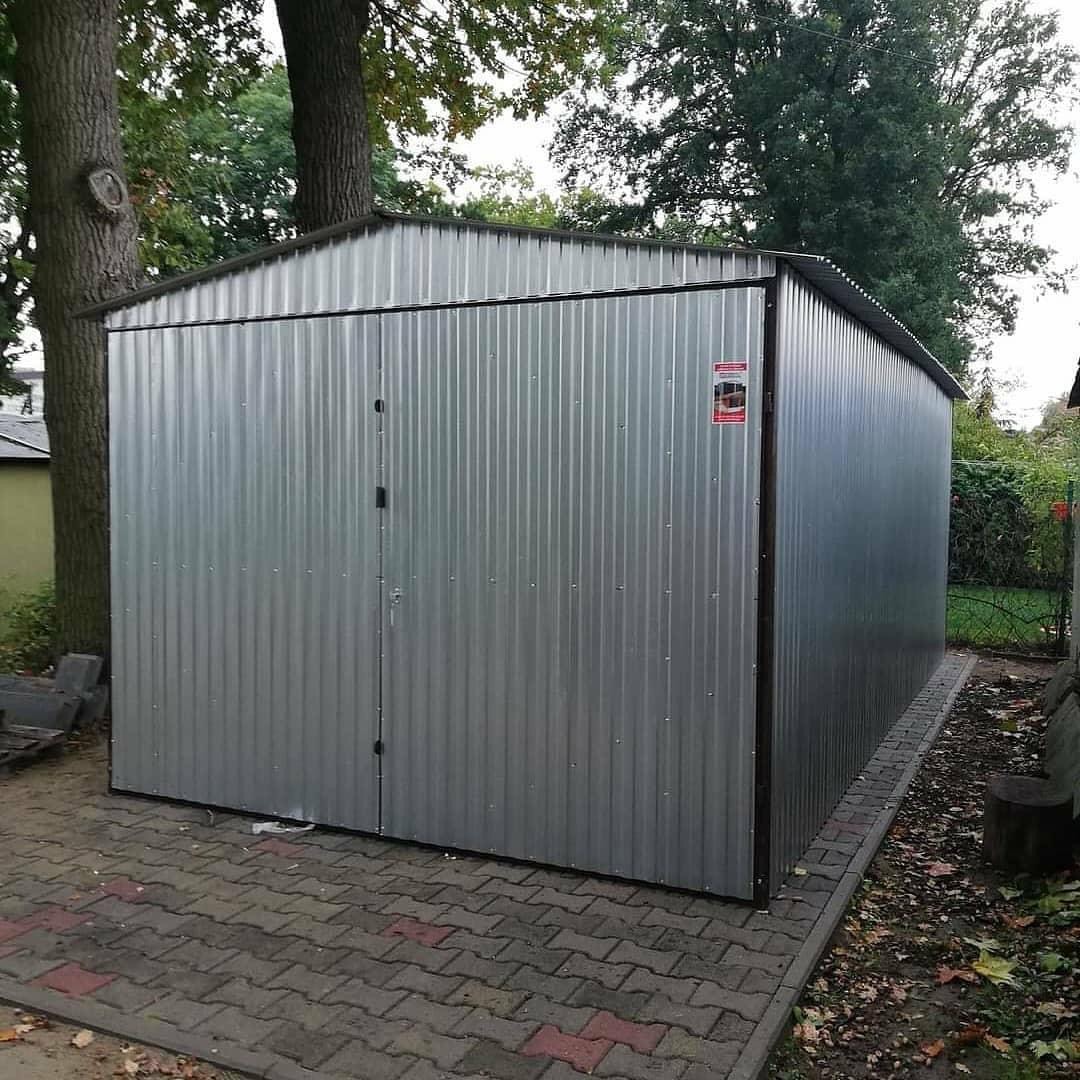 Nowy, srebrny garaż blaszany z dwuskrzydłowymi drzwiami, ustawiony na kostce brukowej w ogrodzie z drzewami w tle.