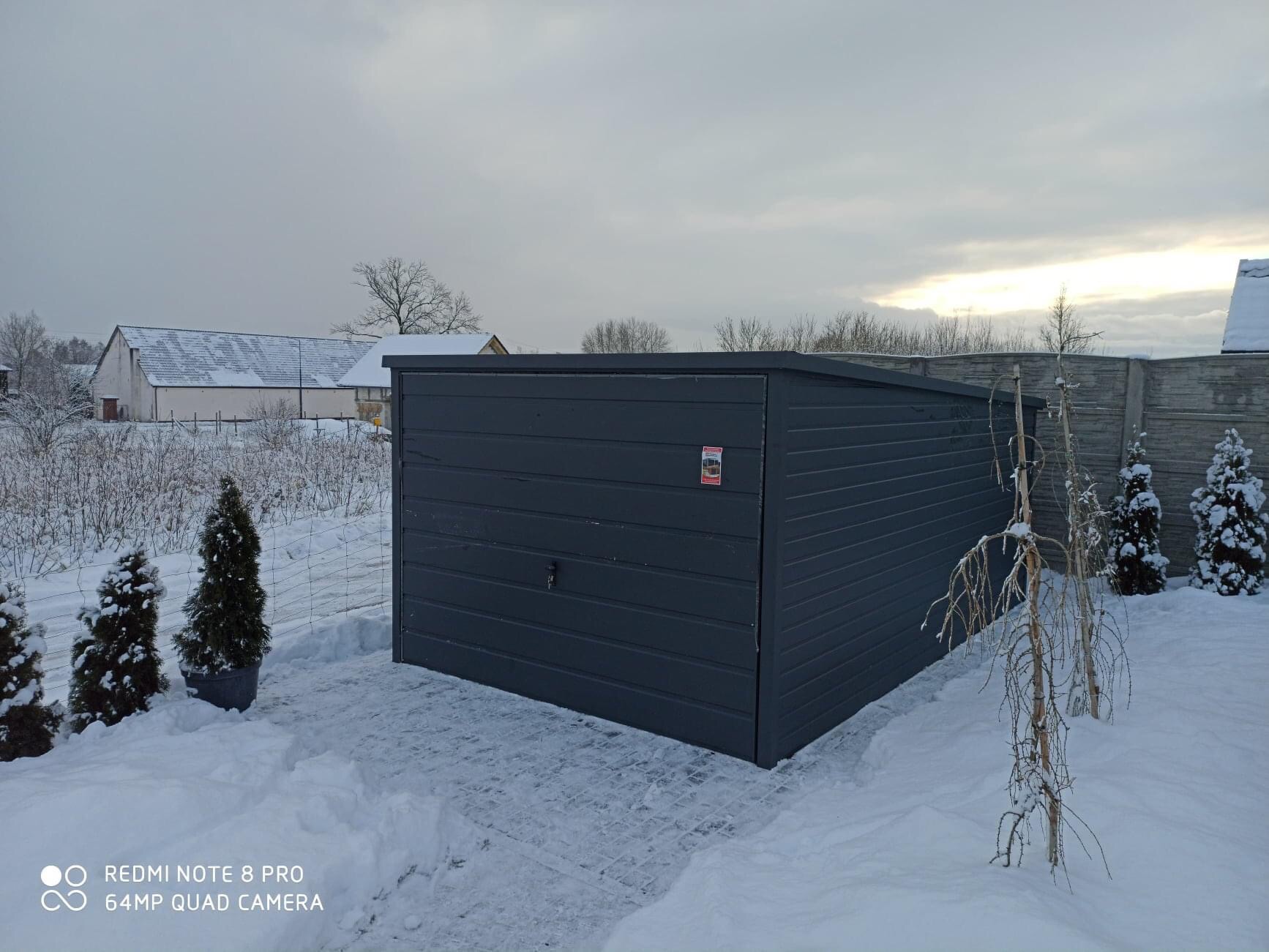 Grafitowy garaż z blachy falistej na tle zimowego krajobrazu, z małymi drzewkami iglastymi i ośnieżonymi budynkami w oddali.
