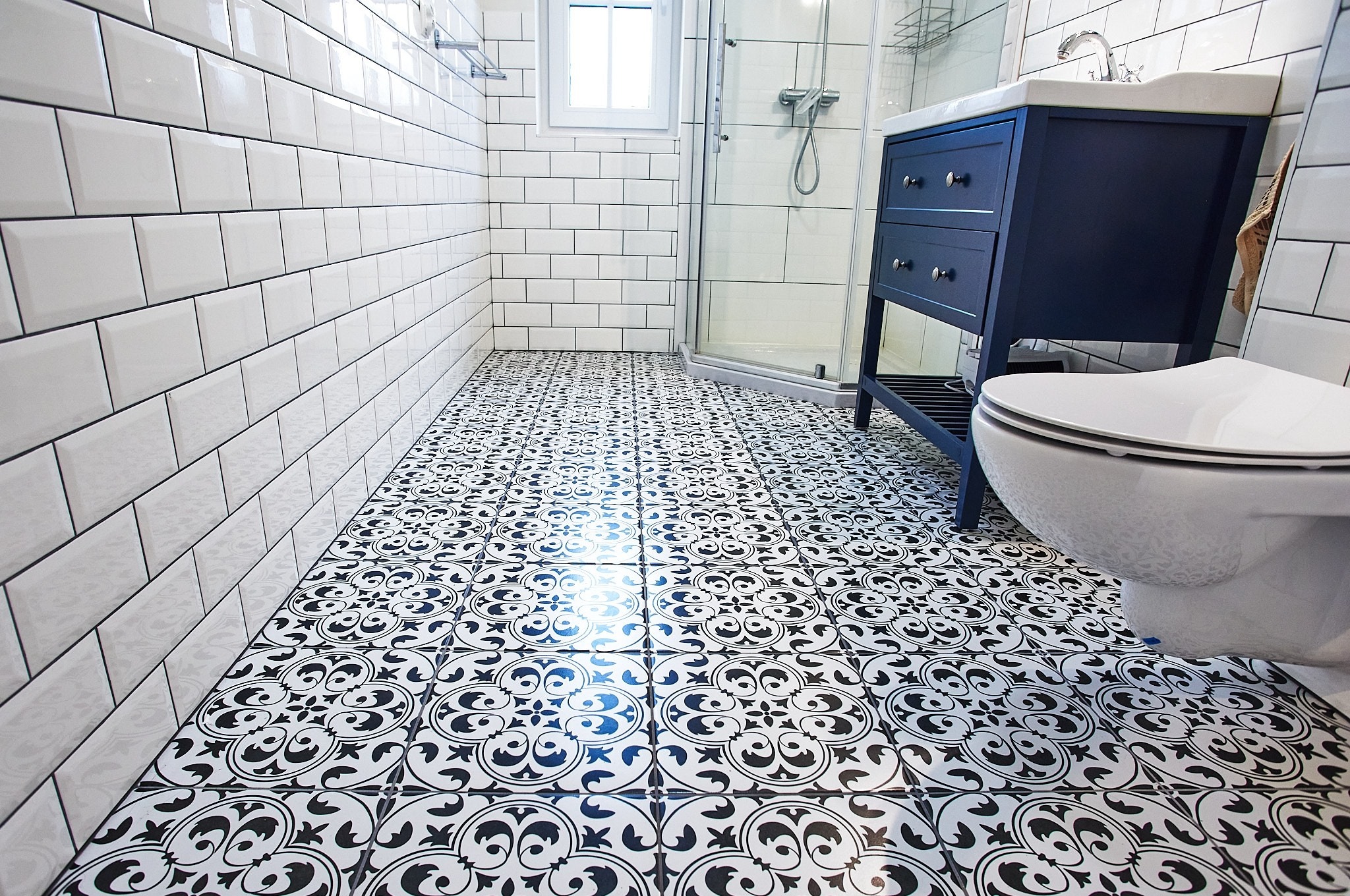 Biała łazienka z czarno-białą wzorzystą podłogą, białe kafelki na ścianach ułożone w cegiełkę, granatowa szafka pod umywalką i kabina prysznicowa w tle.