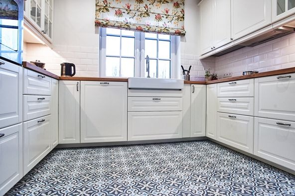 Jasna kuchnia w stylu klasycznym z białymi szafkami, drewnianym blatem i wzorzystą podłogą. Okno z roletą w kwiaty nad ceramicznym zlewem.