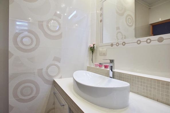 Biała, owalna umywalka nablatowa na białym blacie w nowoczesnej łazience z płytkami w geometryczne wzory, chromowana bateria i różowe świeczki.