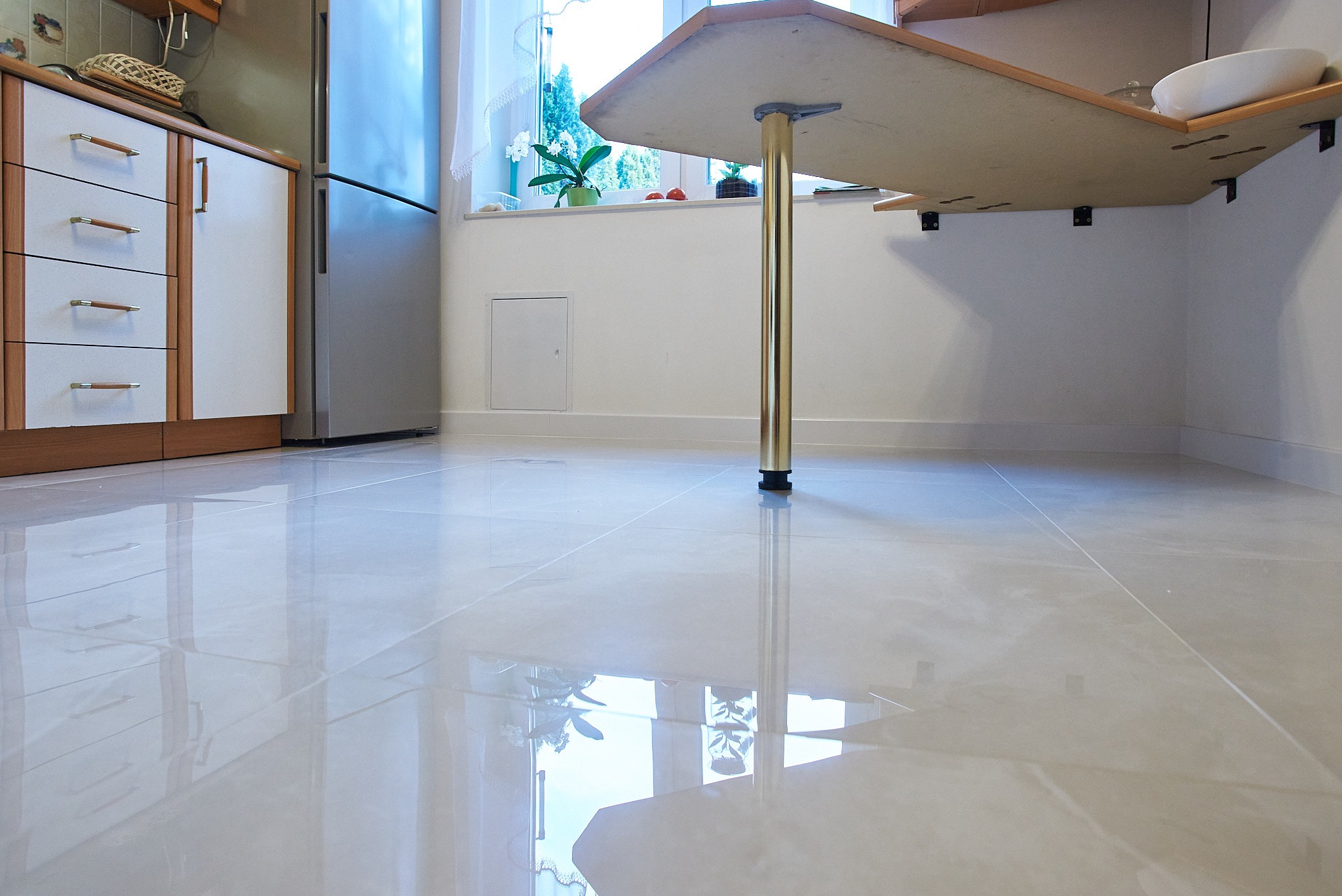 Błyszcząca, jasna podłoga z płytek ceramicznych w kuchni, widoczny fragment zabudowy kuchennej, stół z jedną złotą nogą i odbicie okna z roślinami w podłodze.