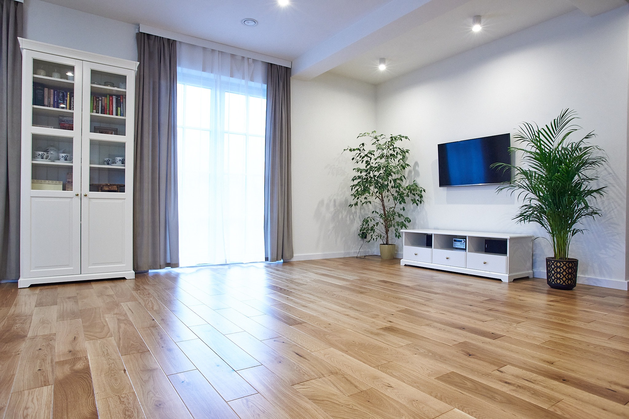 Jasny salon z parkietem ułożonym w jodełkę, białą szafką z przeszklonymi drzwiami, telewizorem na ścianie i roślinami doniczkowymi.