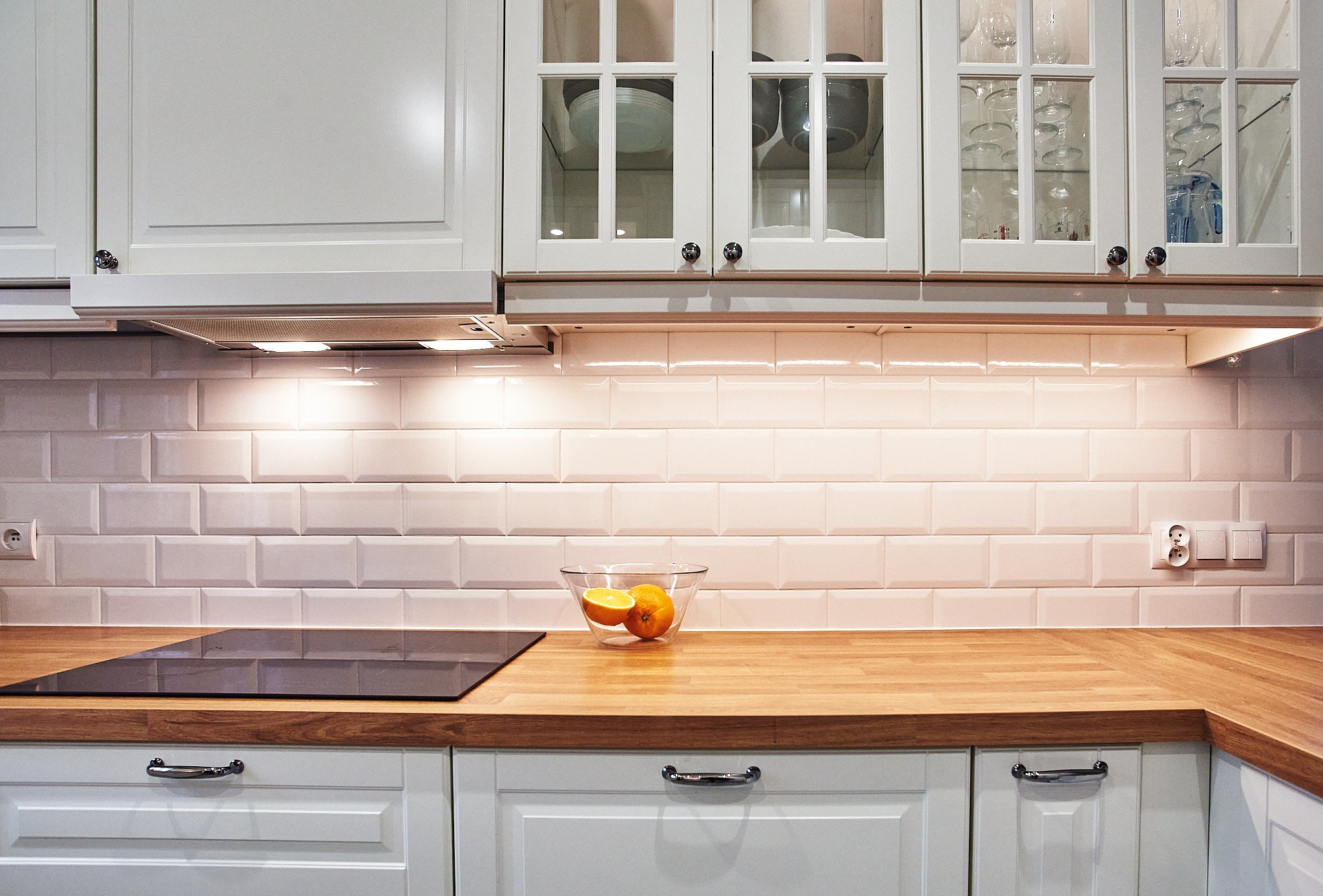 Biała kuchnia z drewnianym blatem, szklanymi szafkami górnymi, podświetlanymi płytkami w stylu metro i owocami w szklanej misce.