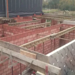 Wznoszenie ścian domu z czerwonej cegły, widoczne szalunki i podpory stropowe, ocieplenie ściany z folią UNILIN, w tle krajobraz.