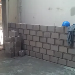 Wznoszenie ściany z szarych bloczków betonowych w surowym wnętrzu, widoczne rusztowanie w tle, poziomica oparta o ścianę, kask i bluza na murze, butelka wody mineralnej.