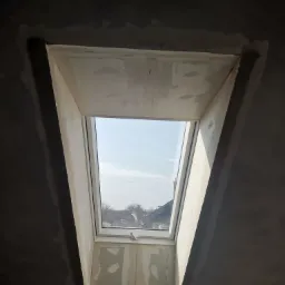 Montaż okna dachowego w surowym wnętrzu, widoczne ślady szpachlowania wokół ramy okiennej i fragment nieotynkowanego sufitu.