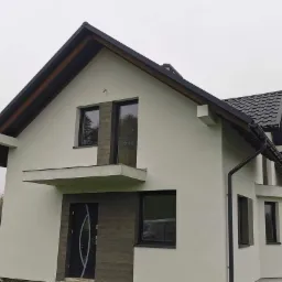 Dwukondygnacyjny dom z jasną elewacją i ciemną stolarką okienną, widok z zewnątrz w pochmurny dzień, widoczny fragment dachu sąsiedniego budynku.