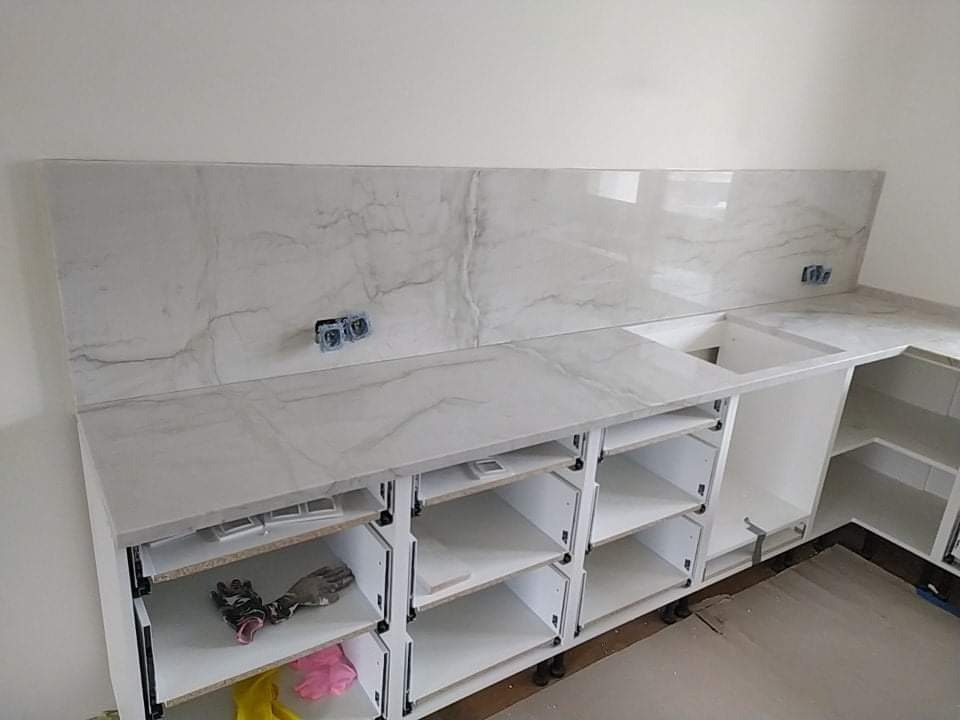 Montaż białej, marmurkowanej płyty na ścianie i blacie nowej, białej zabudowy kuchennej; widoczne gniazdka elektryczne, puste półki i narzędzia w szufladach.