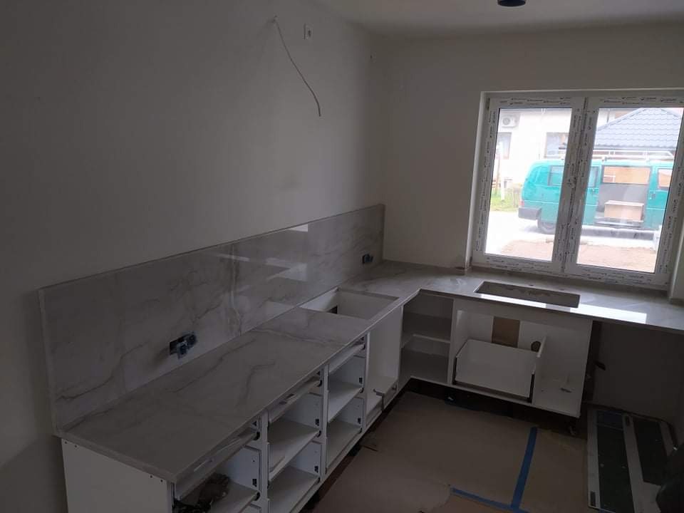 Wnętrze kuchni w trakcie montażu: białe szafki bez frontów, marmurowe blaty i panel ścienny, widoczne okno z zielonym busem na zewnątrz, podłoga zabezpieczona kartonem.