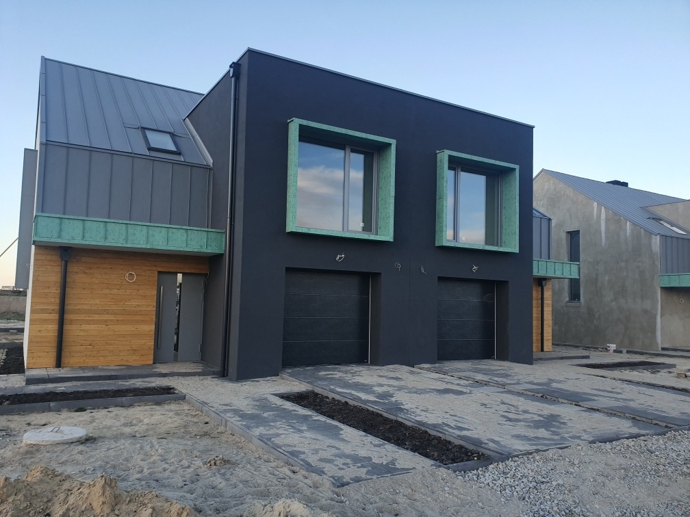 Nowoczesny bliźniak z ciemną elewacją, drewnianą okładziną, szarą blachą na dachu i zielonymi obramowaniami okien, na etapie wykończenia terenu.