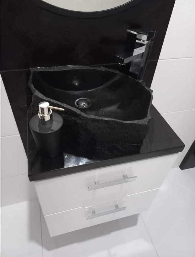 Czarna, kamienna umywalka o nieregularnym kształcie na białej szafce z dwoma szufladami i chromowanymi uchwytami, z czarnym blatem i baterią w tle, uzupełniona czarnym dozownikiem mydła w łazience...