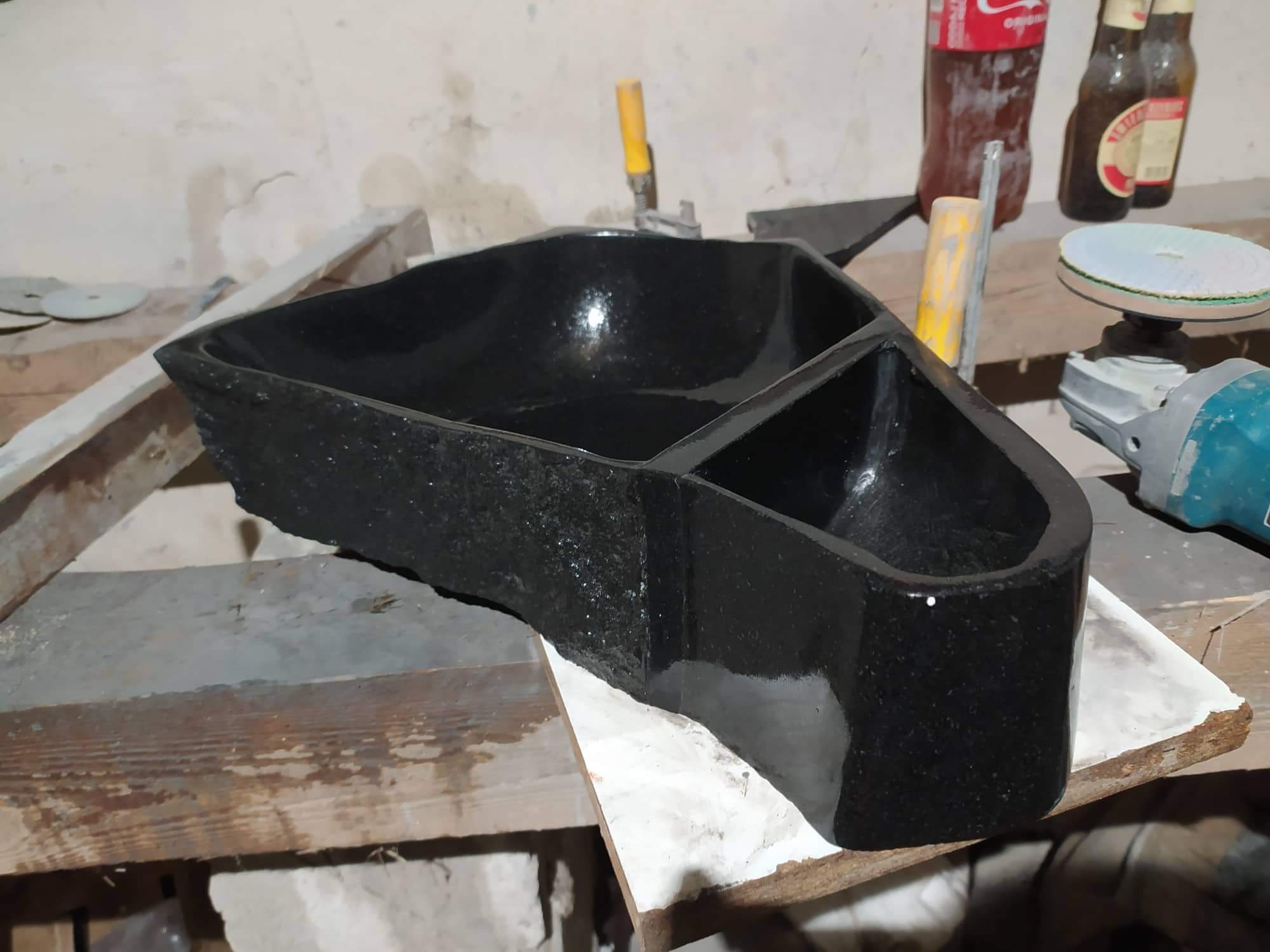 Czarna, nietypowa umywalka z kamienia, podzielona na dwie części, stoi na drewnianym stole warsztatowym, obok narzędzia i napoje.