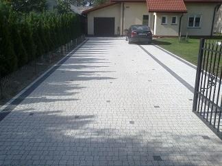 Ułożona kostka brukowa na podjeździe do domu z garażem, widok zza otwartej bramy wjazdowej, cień drzew na nawierzchni, zadbany trawnik w tle.