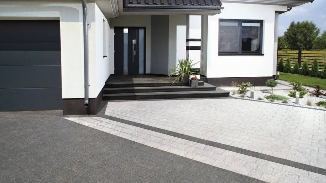 Nowoczesny dom jednorodzinny z elewacją w kolorze białym i szarym, czarne okna i drzwi, podjazd wyłożony kostką brukową w dwóch odcieniach szarości, minimalistyczny ogród z niskimi krzewami i lampami.