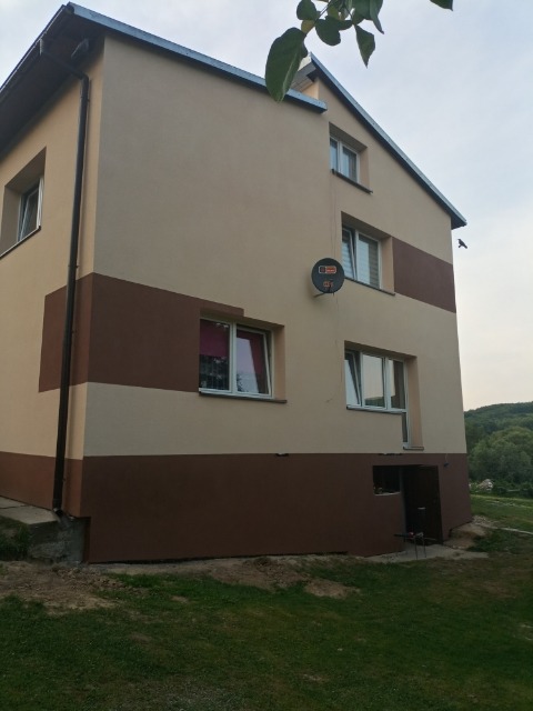 Dwukondygnacyjny dom z elewacją w odcieniach beżu i brązu, z widoczną anteną satelitarną, oknami z białymi ramami i zielonym otoczeniem w tle.