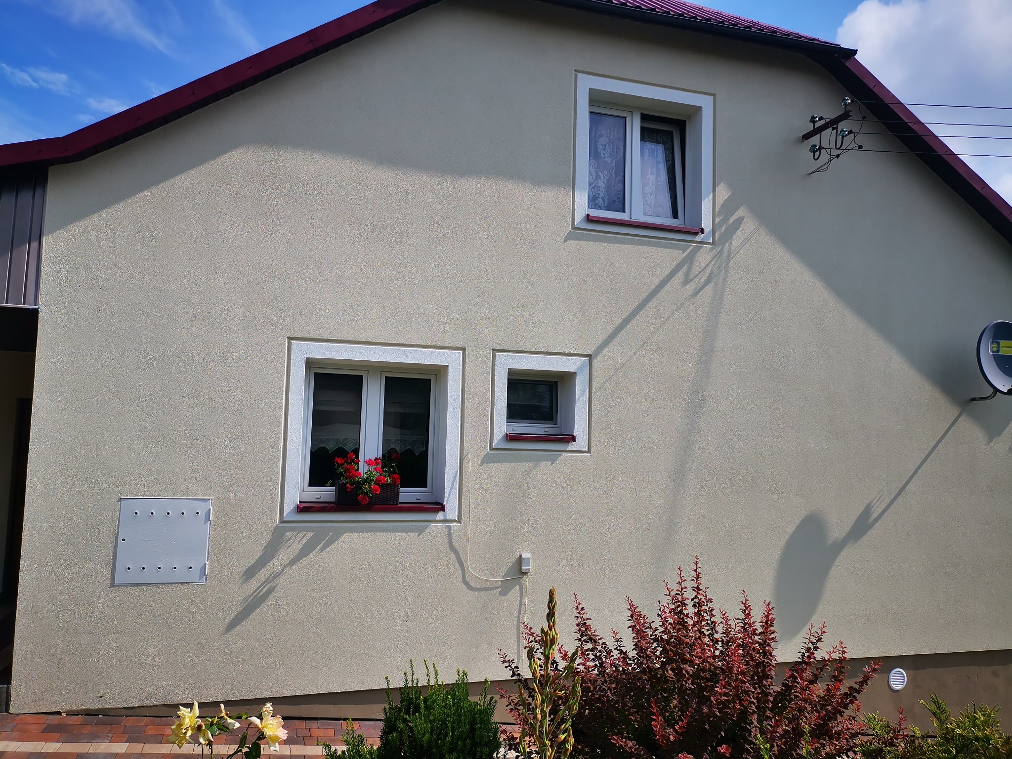 Jasna elewacja domu jednorodzinnego z trzema oknami o różnych rozmiarach, z kwiatami w doniczce na parapecie jednego z okien, widoczne przyłącze elektryczne i antena satelitarna.