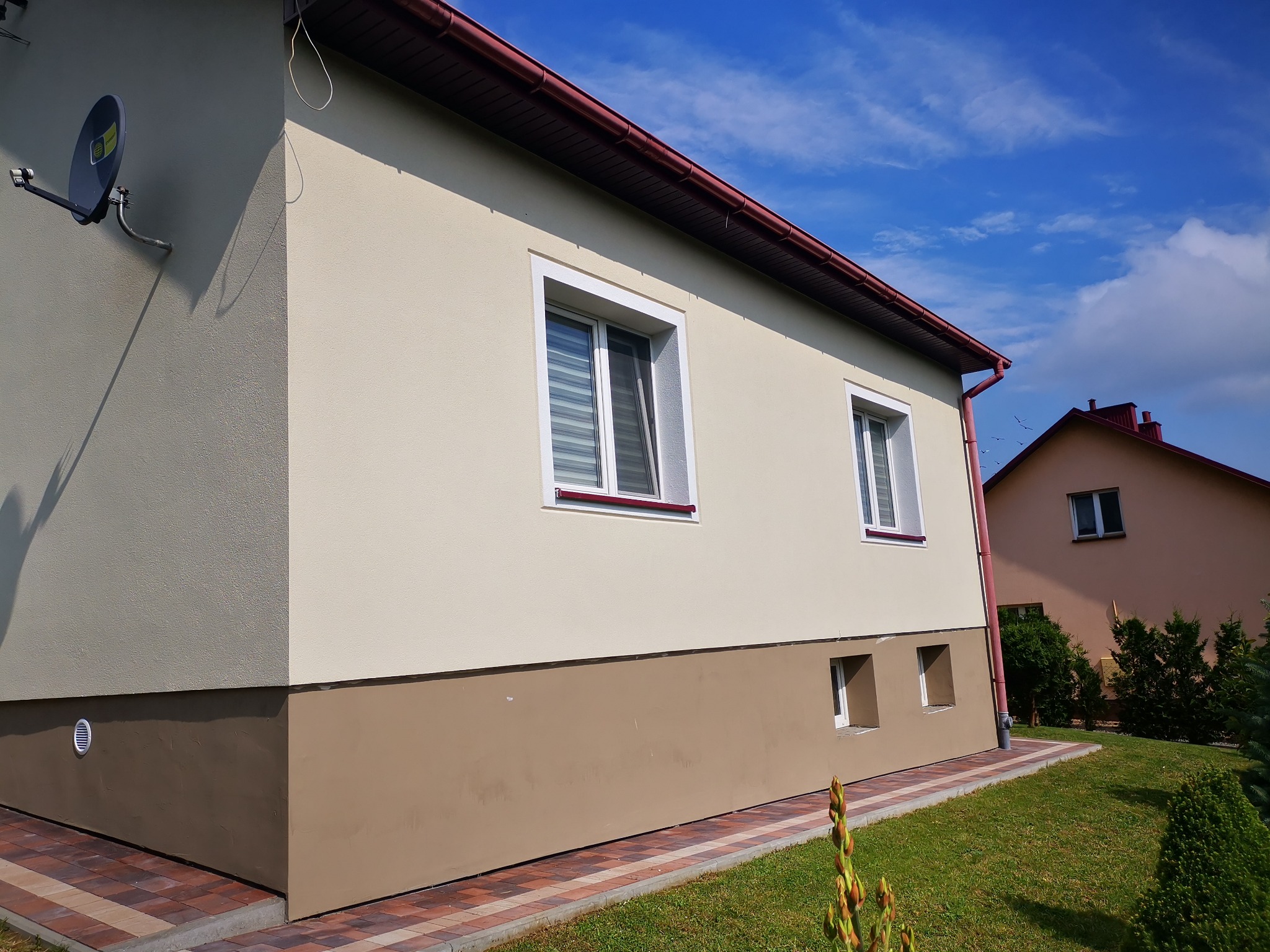 Ocieplony dom jednorodzinny z beżową elewacją i brązowym cokołem, widoczne dwa okna z roletami, antena satelitarna na ścianie szczytowej, czerwona rynna i kostka brukowa wokół budynku.