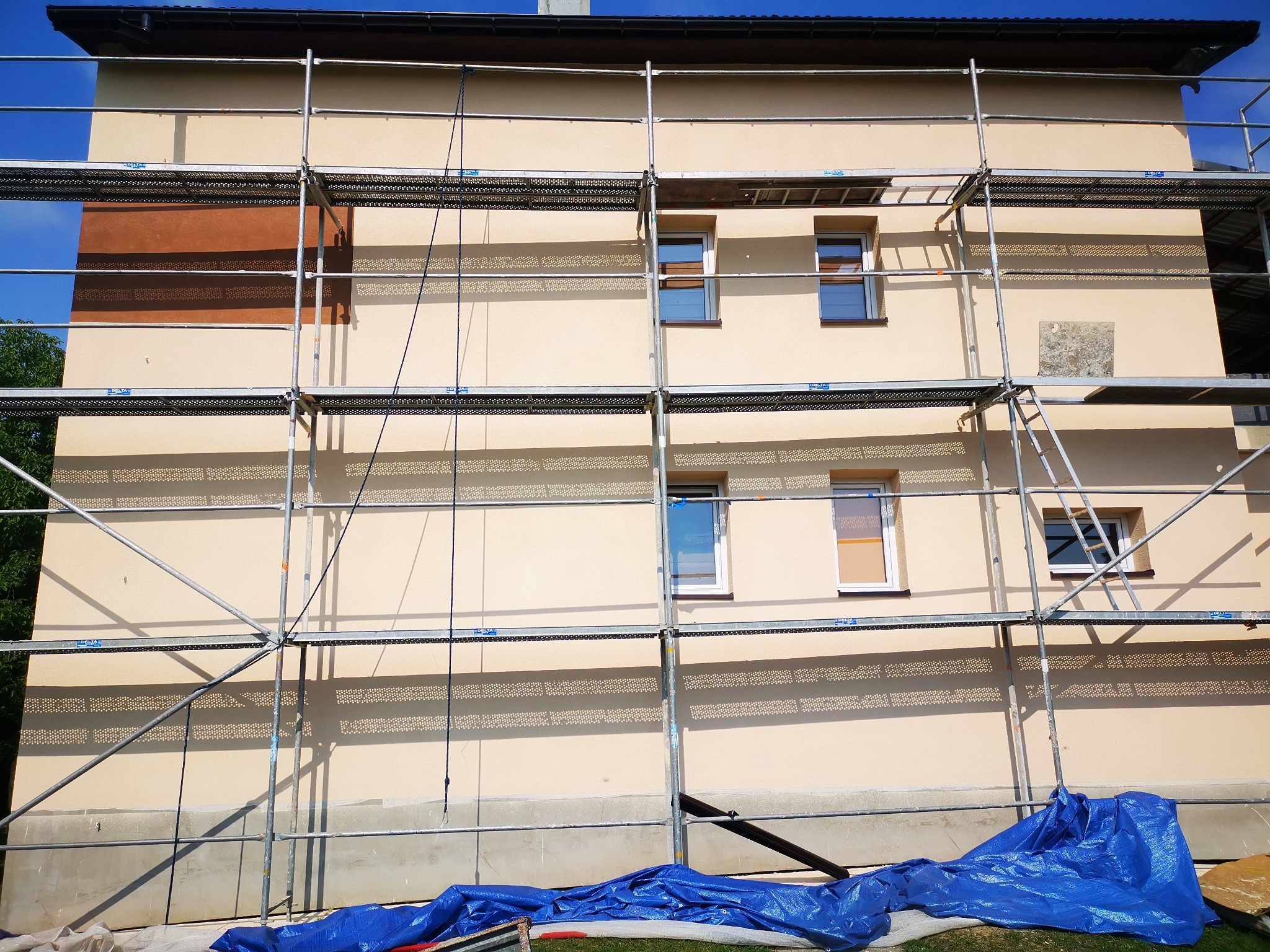 Dom w trakcie prac elewacyjnych z widocznym rusztowaniem, fragmentem ocieplenia w kolorze brązowym i zabezpieczającą folią w kolorze niebieskim u podstawy ściany.