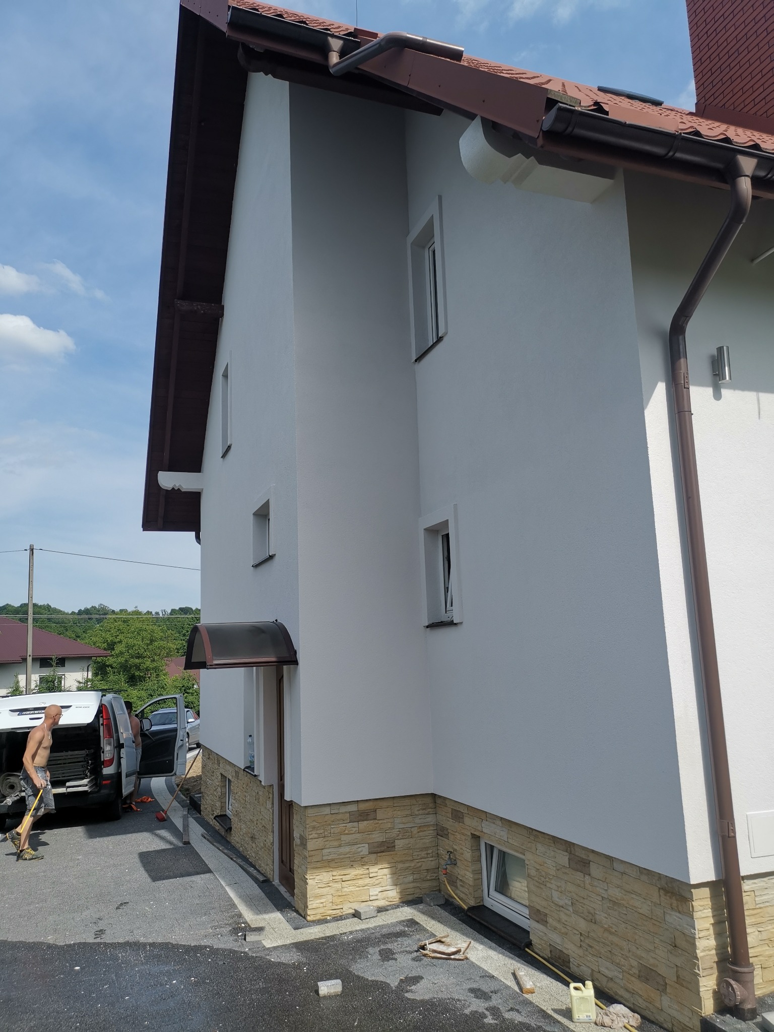 Świeżo otynkowany dom jednorodzinny z brązowym dachem i kamienną podmurówką, widoczny pracownik przy samochodzie dostawczym i mokry chodnik.