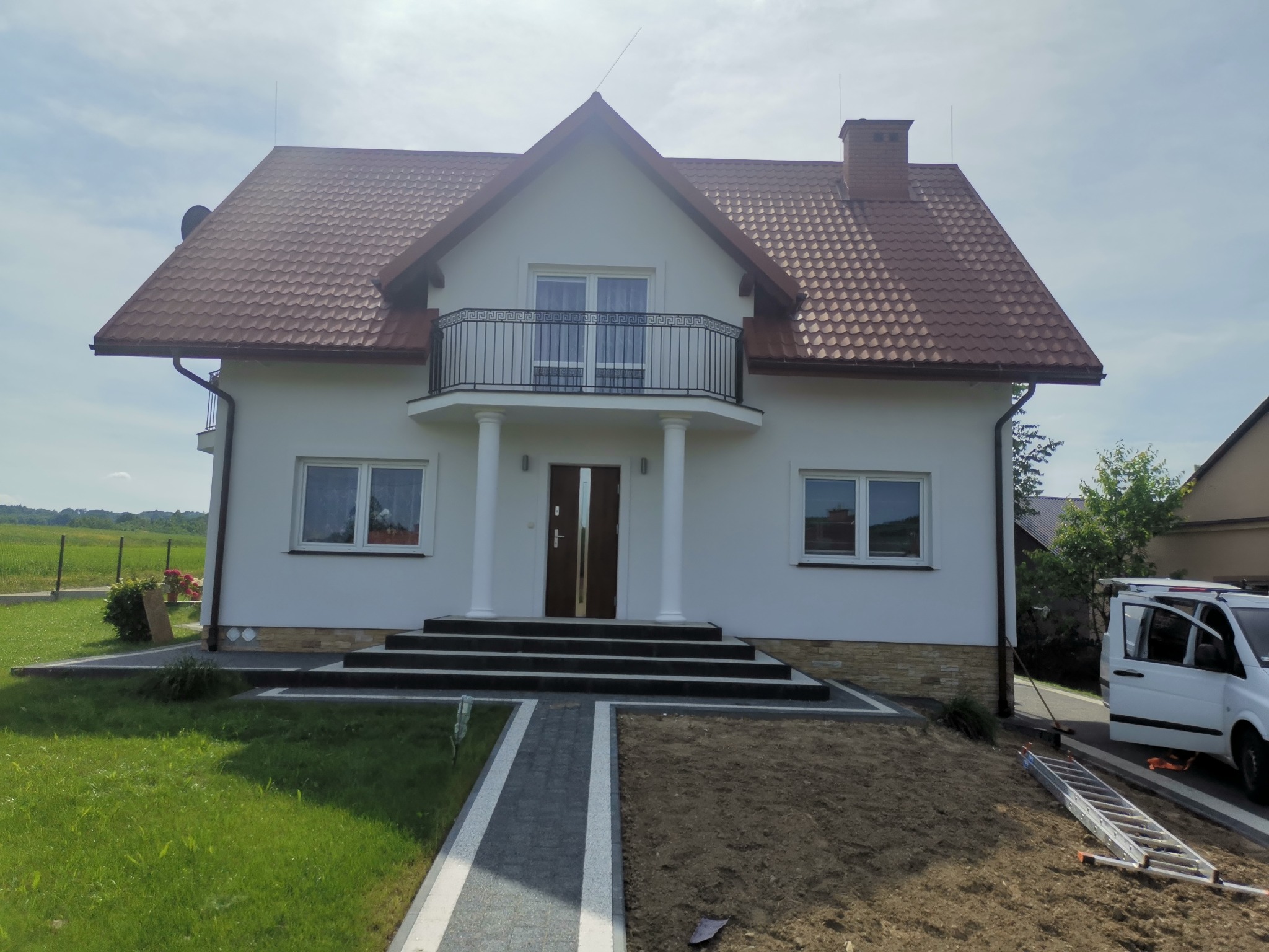 Biały dom jednorodzinny z brązowym dachem, balkonem z ozdobną balustradą i kolumnami przy wejściu, widok z przodu, z fragmentem terenu przed domem, gdzie widać częściowo rozkopaną ziemię,...